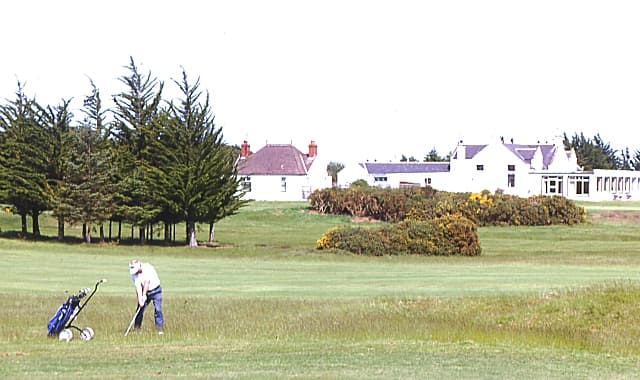 A golfer in the rough at Elgin, the clubhouse beyond the gorse